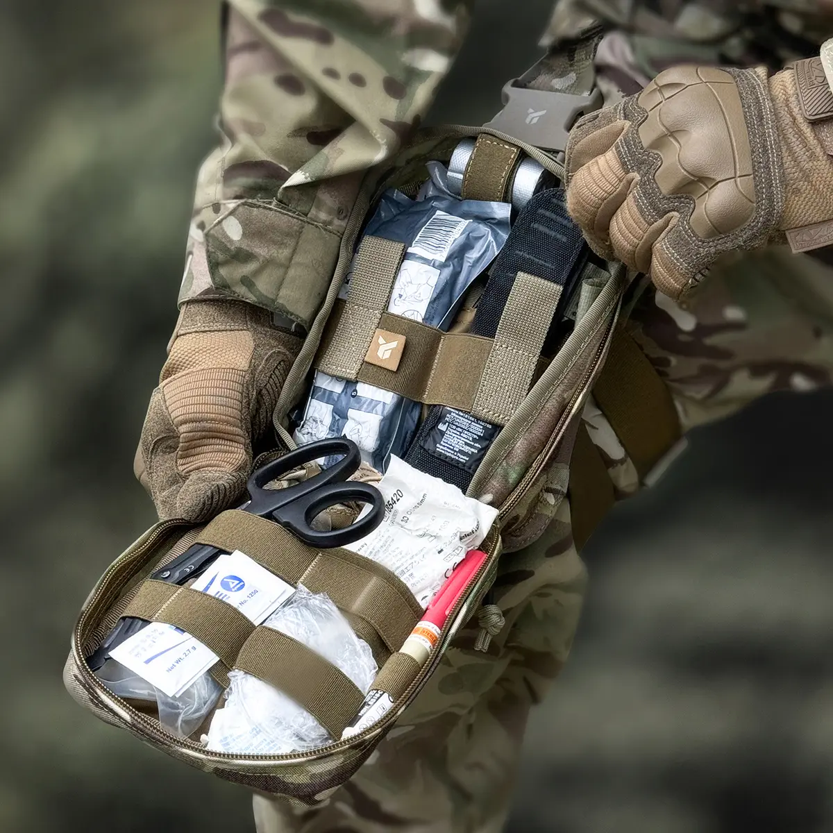 Tasca medica cosciale PM21 Multicam aperta in ambiente operativo, organizzata con bende compressive, forbici da trauma e laccio emostatico.