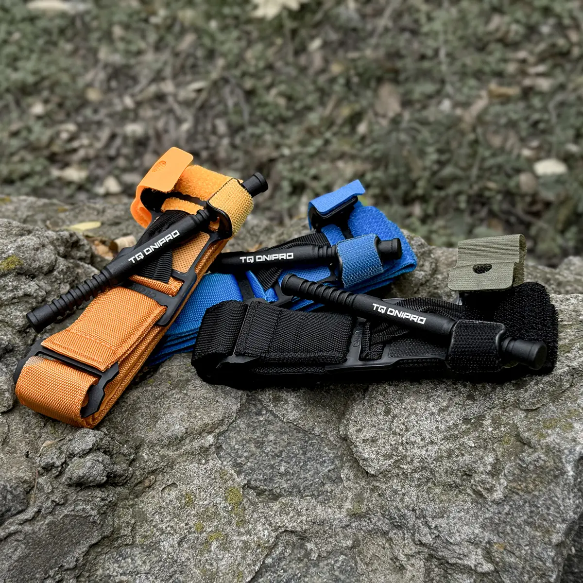 Tourniquet TQ Dnipro arancione con versioni blu e nera appoggiati su roccia per confronto.